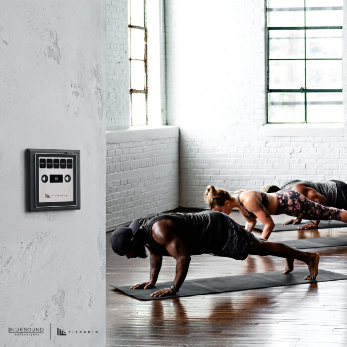 A man and a woman are working out inside a gym and a bluesound professional CP200 music controller on wall for music in the gym